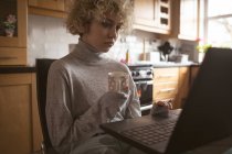 Jovem mulher usando laptop enquanto toma café em casa — Fotografia de Stock