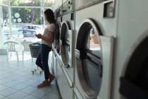 Giovane donna che utilizza il suo telefono in attesa di lavanderia — Foto stock