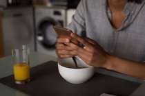 Mulher usando telefone celular enquanto toma café da manhã em casa — Fotografia de Stock