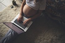 Mujer embarazada usando el ordenador portátil en la sala de estar en casa - foto de stock