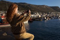 Mulher tirando foto do porto com telefone celular à luz do sol — Fotografia de Stock