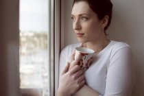 Mulher cuidadosa segurando caneca de café e olhando através da janela . — Fotografia de Stock