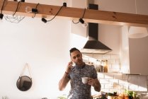 Чоловік з чашкою кави розмовляє на мобільному телефоні на кухні вдома . — стокове фото