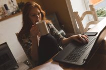 Жінка має каву під час використання ноутбука вдома — стокове фото