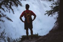 Rückansicht eines Mannes, der mit den Händen an der Hüfte in der Nähe eines Wasserfalls steht — Stockfoto