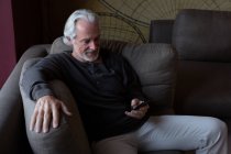 Homem sênior usando telefone celular na sala de estar em casa — Fotografia de Stock