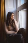 Задумчивая женщина смотрит в окно, сидя дома на подоконнике — стоковое фото