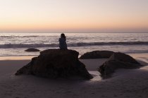 Silhouette einer Frau, die sich in der Abenddämmerung auf einem Felsen am Strand entspannt. — Stockfoto