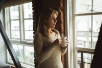 Продумана жінка дивиться крізь вікно, маючи каву вдома — стокове фото