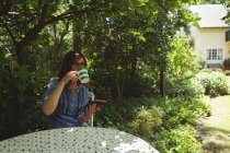 Задумчивая женщина пьет кофе во время использования цифрового планшета в саду — стоковое фото