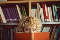Porträt einer jungen Frau, die ihr Gesicht hinter Buch in Bibliothek versteckt — Stockfoto