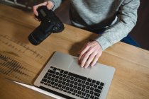 Homem usando laptop enquanto segurando câmera digital em casa — Fotografia de Stock
