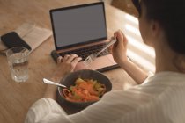 Молода жінка їсть сніданок, дивлячись в ноутбук у вітальні вдома — стокове фото