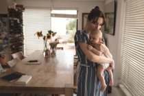 Young mom in bath rob holding and kissing her baby in living room at home — Stock Photo