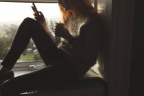 Mulher usando telefone celular enquanto toma café em casa — Fotografia de Stock