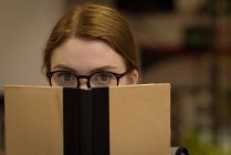 Nahaufnahme einer Frau mit Brille, die ihr Gesicht hinter einem Buch in der Bibliothek versteckt — Stockfoto