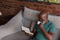 Homme âgé prenant un café tout en lisant un livre à la maison — Photo de stock