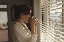 Jovem mulher de pé perto da janela tomando café em casa — Fotografia de Stock