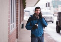 Молодой человек разговаривает по мобильному телефону во время прогулки по улице с кофе . — стоковое фото