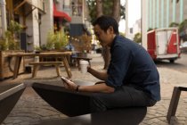 Улыбающийся азиатский бизнесмен с помощью мобильного телефона в тротуарном кафе — стоковое фото