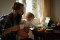 Pai tomando café enquanto sol jogando no laptop em casa — Fotografia de Stock