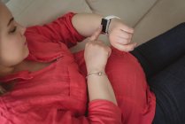 Primo piano della donna incinta seduta sul divano a controllare il suo orologio intelligente a casa — Foto stock
