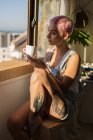 Stylische Frau mit rosa Haaren, die zu Hause Kaffee und Handy im Sonnenlicht hält. — Stockfoto