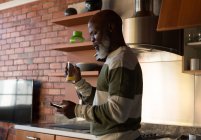 Старший мужчина пьет сок дома, пользуясь мобильным телефоном — стоковое фото