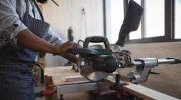 Sección media del carpintero cortando tablón de madera con sierra eléctrica en taller - foto de stock