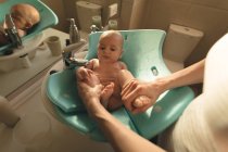 Mother washing a baby in baby bath seat in bathroom sink at home — Stock Photo