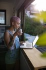 Junge Frau mit rosa Haaren trinkt Kaffee, während sie Laptop in der Nähe des Fensters zu Hause benutzt. — Stockfoto