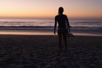 Силуэт подходящей женщины, держащей доску для серфинга на пляже в сумерках . — стоковое фото