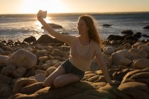 Mujer sonriente tomando selfie en la playa al atardecer - foto de stock