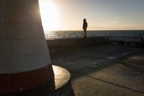 Femme observant le paysage marin près du phare pendant le coucher du soleil — Photo de stock