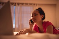 Женщина разговаривает по мобильному телефону во время использования ноутбука в спальне дома — стоковое фото