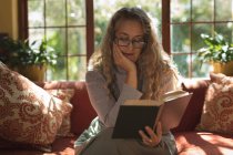Reife Frau sitzt auf Sofa, während sie zu Hause Roman liest — Stockfoto