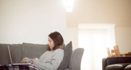 Mujer sosteniendo la taza de café mientras usa el ordenador portátil en el sofá en la sala de estar en casa
. - foto de stock
