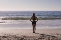 Fit mulher de pé com as mãos sobre os quadris na praia ao entardecer, visão traseira . — Fotografia de Stock