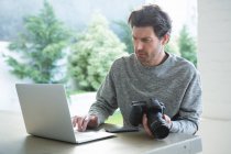 Homem usando laptop e segurando câmera digital em casa — Fotografia de Stock