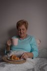 Senior mulher tomando café e café da manhã no quarto em casa — Fotografia de Stock