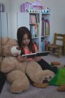 Criança idade elementar com ursinho de pelúcia leitura livro na sala de estar em casa — Fotografia de Stock