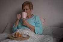 Пожилая женщина с кофе и завтраком в спальне дома — стоковое фото