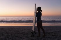 Silhouette di donna in forma che tiene la tavola da surf in spiaggia al tramonto . — Foto stock