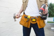 Mid section of male carpenter with tool belt holding hammer and drill machine — Stock Photo