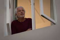 Homem idoso atencioso olhando através da janela em casa — Fotografia de Stock