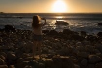 Женщина фотографирует море с помощью мобильного телефона на закате — стоковое фото