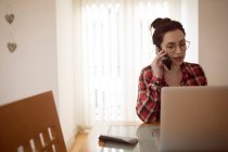 Woman talking on mobile phone while using laptop at home. — Stock Photo