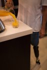 Vista cortada da mulher com prótese perna derramando suco de laranja em vidro em casa . — Fotografia de Stock