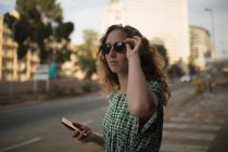 Thoughtful woman in sunglasses standing on roadside, holding smartphone — Stock Photo