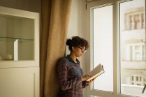 Donna che legge libro vicino alla finestra a casa — Foto stock